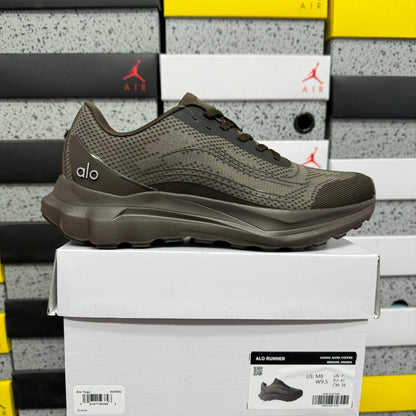 A dark olive green Alo Runner sneaker is displayed on top of its white shoebox, with stacked Jordan shoeboxes in the background.